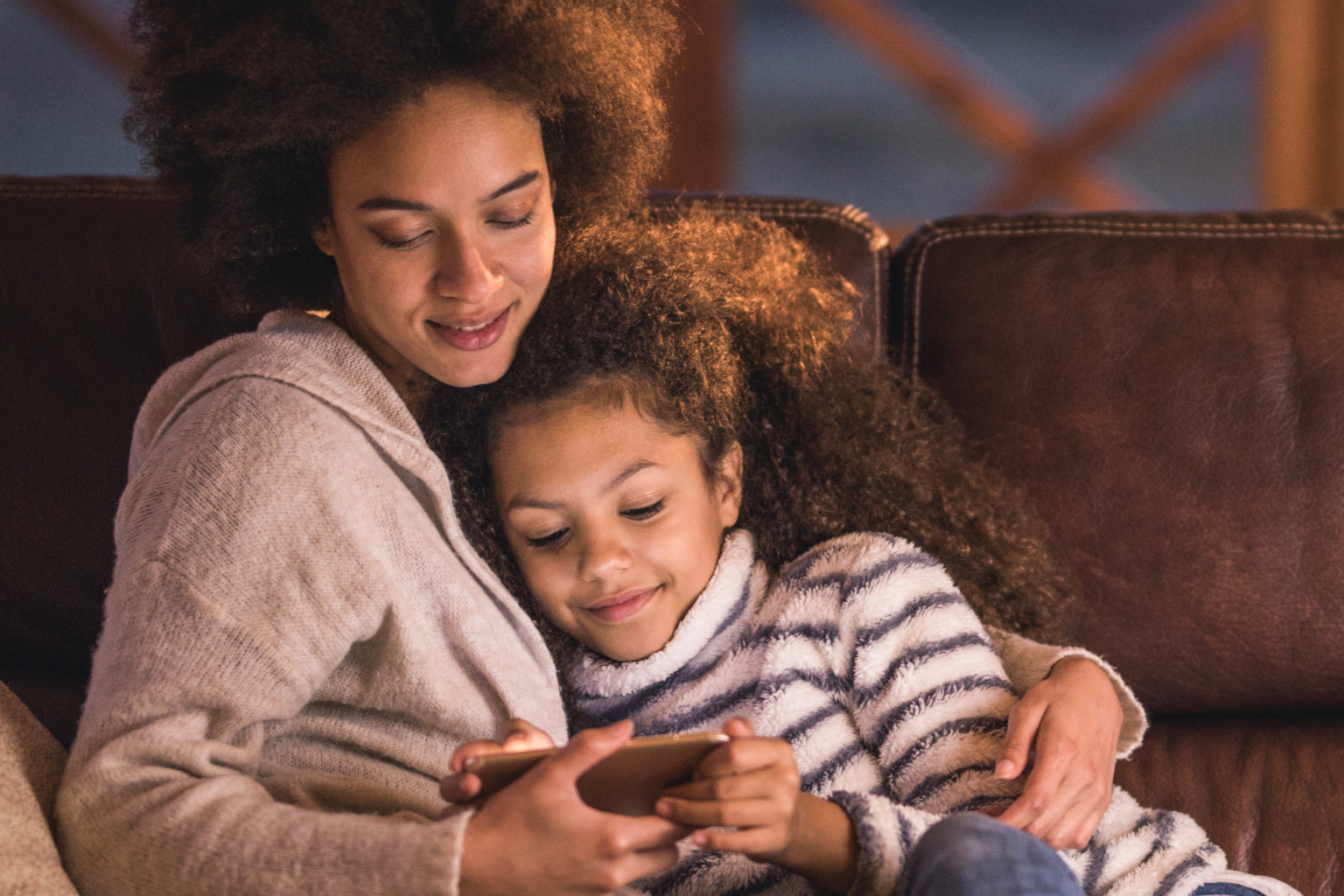 A mother and daughter cuddling on a couch while looking at a phone.